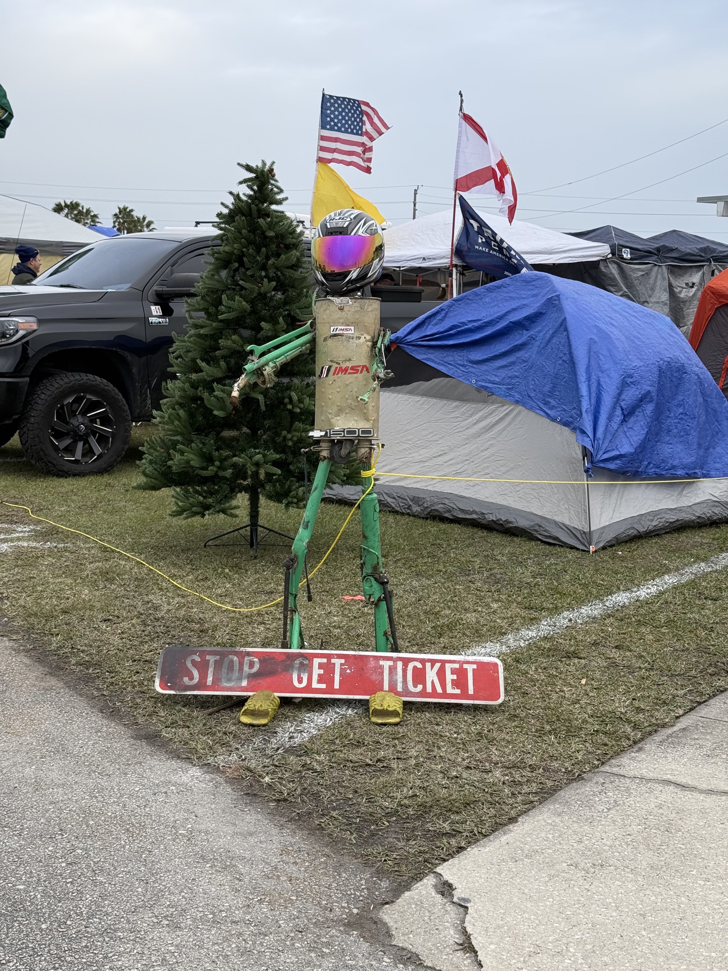 A strange folk art statue in front of a car camping site in the daytime